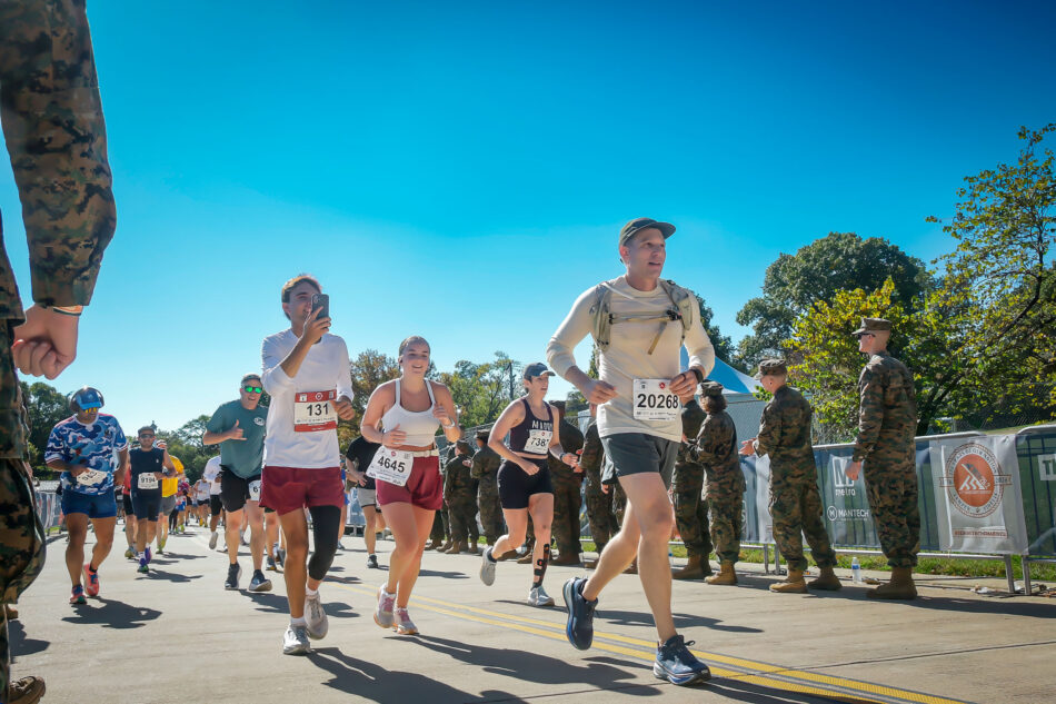 Devon Akmon, wearing bib 20268, a cap, a light long-sleeve shirt and a hydration vest, strides along a sunny road during the 2026 Marine Corps Marathon. Other runners follow behind him, and Marines in camouflage uniforms cheer from the barricades under a clear blue sky.