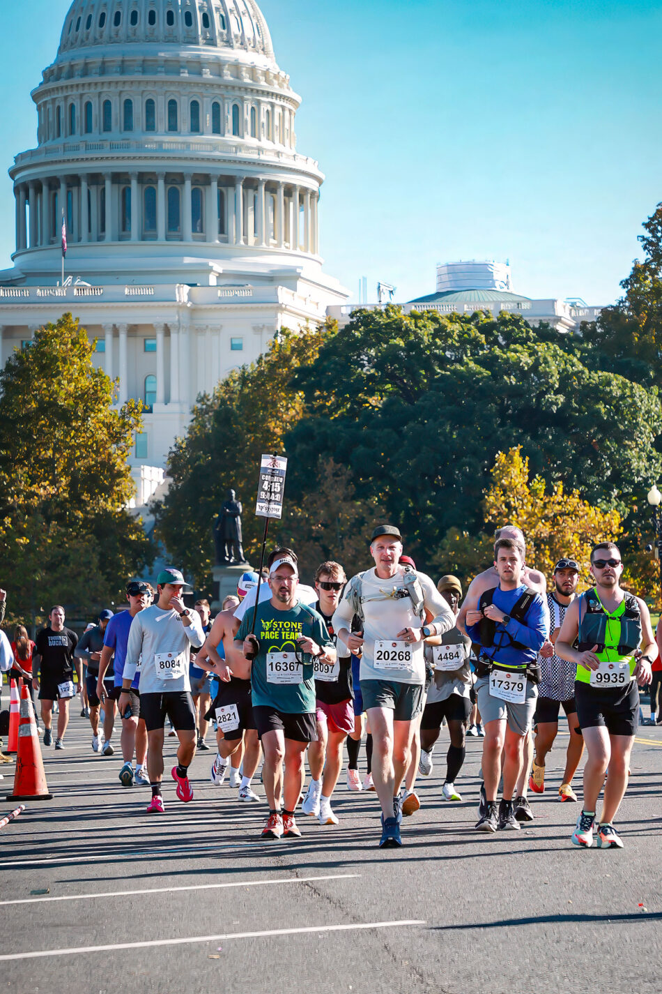 Devon Akmon, wearing bib 20268, runs down a confetti-covered street in Georgetown during the 2026 Marine Corps Marathon, surrounded by other runners. Historic brick buildings and a gold-domed bank building line the street behind them.
