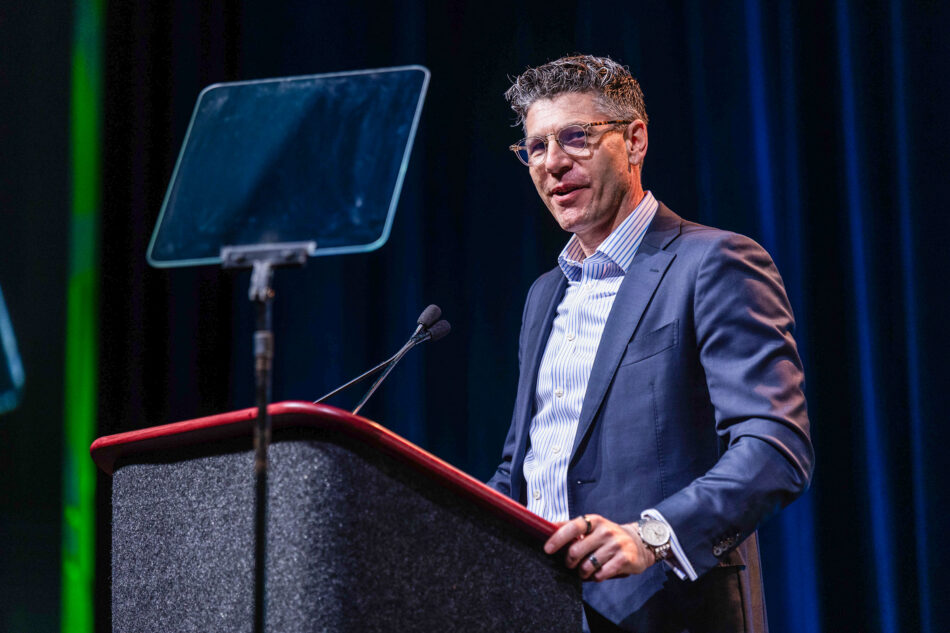 A man in a navy blazer and striped dress shirt speaks at a podium during a conference general session. A teleprompter is visible to his left against a dark draped backdrop with green accent lighting.