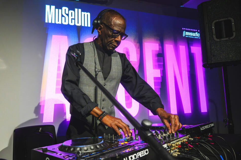 DJ John Collins wearing headphones and glasses, playing music on a modern DJ controller at an event. The background features bright purple lighting and the word 'Museum' in bold white text.