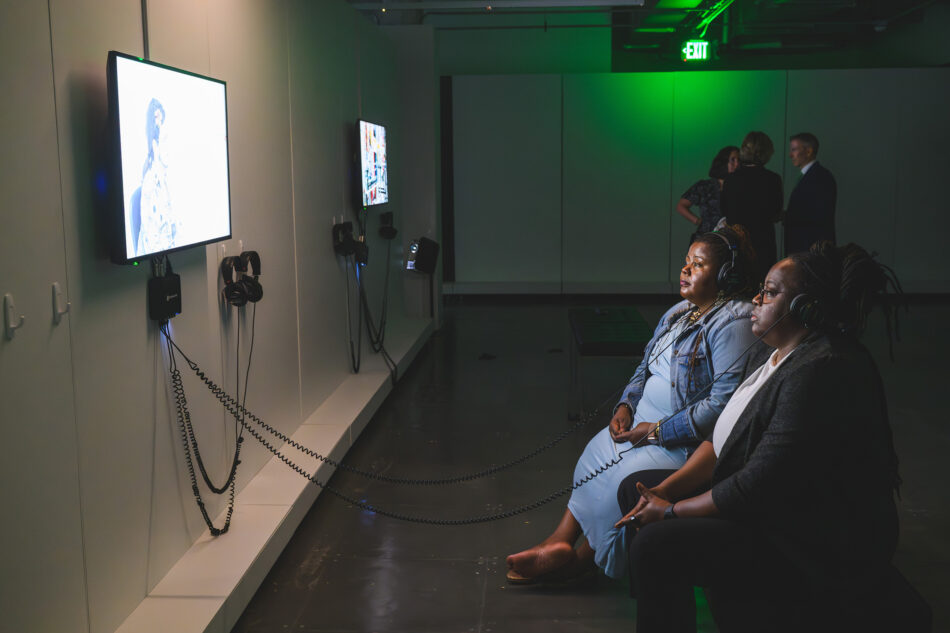 Two women seated on a bench, wearing headphones and watching a video exhibit on a wall-mounted screen in a dimly lit room. Another group of people stands in the background, bathed in green lighting.