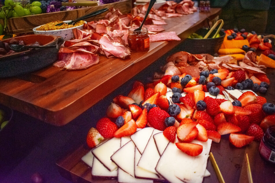 A vibrant food spread featuring sliced strawberries, blueberries, cheese, charcuterie, and condiments arranged on wooden boards.