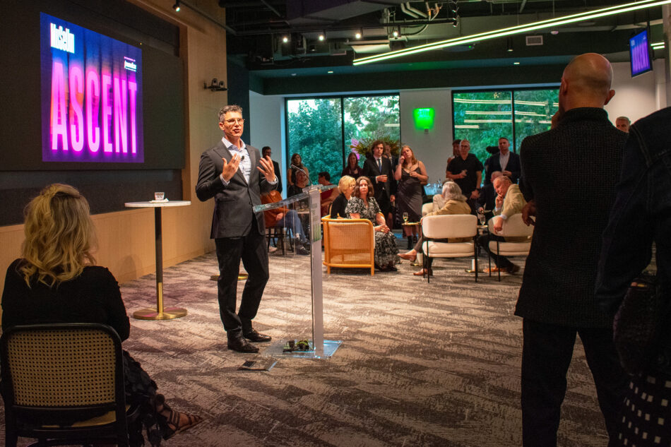 Devon Akmon standing at a podium, addressing an audience at an indoor event. The screen behind displays the word 'Ascent' in large purple letters, and the audience listens intently.