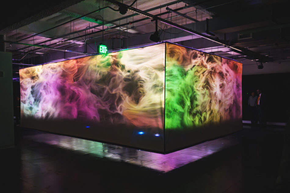 Monumentum by Abel Korinsky appears as a large rectangular art installation with swirling multicolored projections glowing in shades of green, pink, and purple in a dimly lit room.