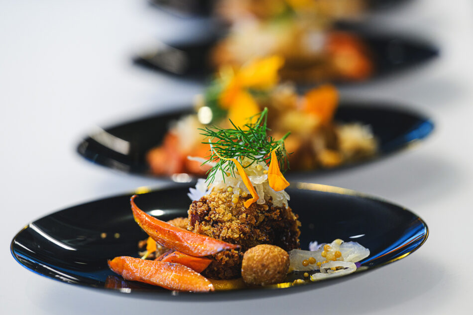 A close-up of elegantly plated gourmet food featuring a small dish garnished with colorful vegetables and herbs, presented on a black plate.