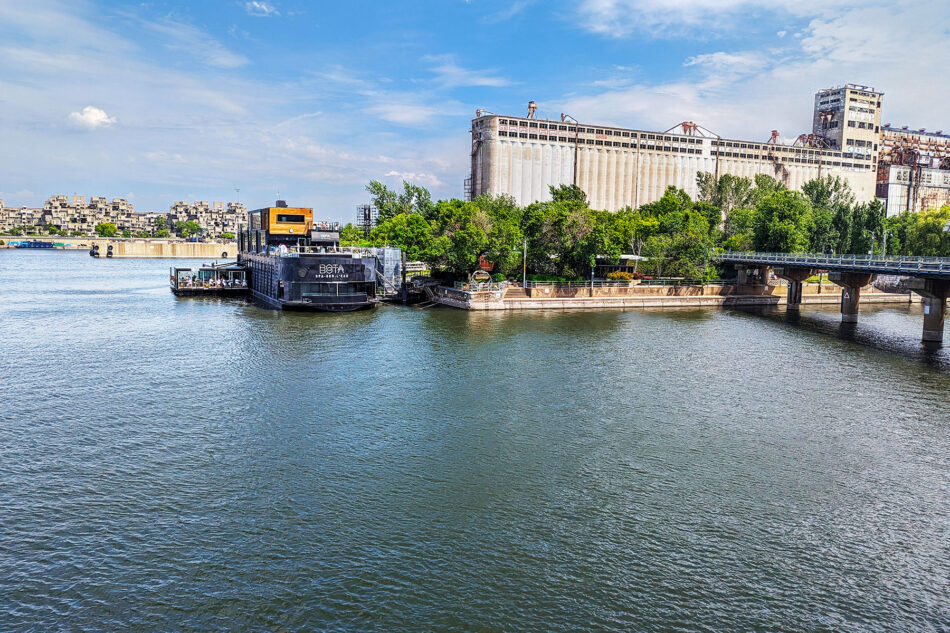 View of Bota Bota spa floating on the Saint Lawrence River in Montreal, with the iconic Habitat 67 residential complex in the background and a large silo structure on the right. The spa features open decks and lush greenery, nestled against the urban backdrop of Montreal's cityscape.