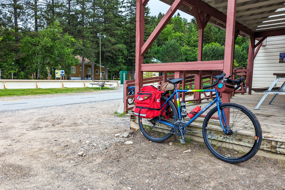 Loaded bicycle leaning against a wooden structure at a rest area along the P'tit Train du Nord trail, with trees and a picnic table in the background.