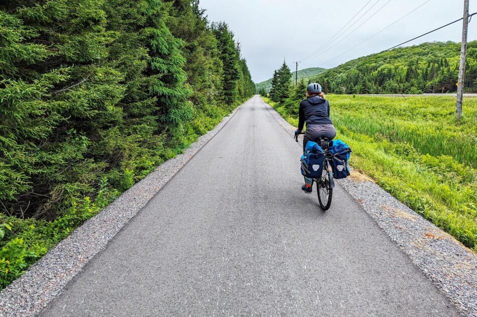 Dharma riding on a paved section of the P'tit Train du Nord trail, with dense greenery on one side and open fields on the other.