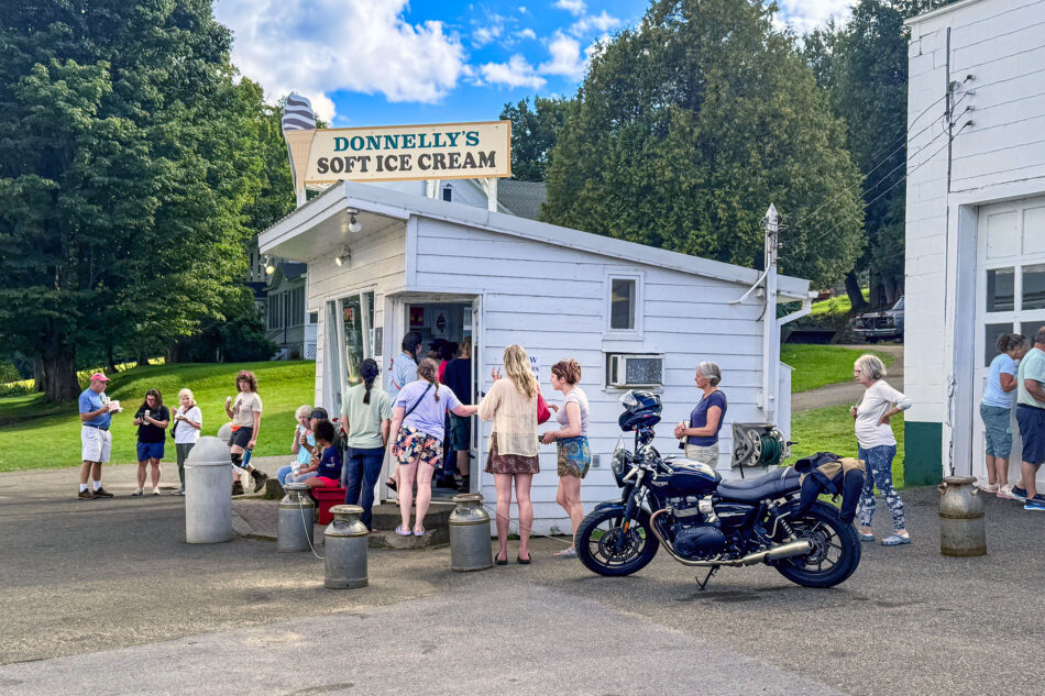 A crowd of people wait outside a small white roadside stand topped with a 'Donnelly's Soft Ice Cream' sign, with a parked motorcycle nearby on a sunny day.