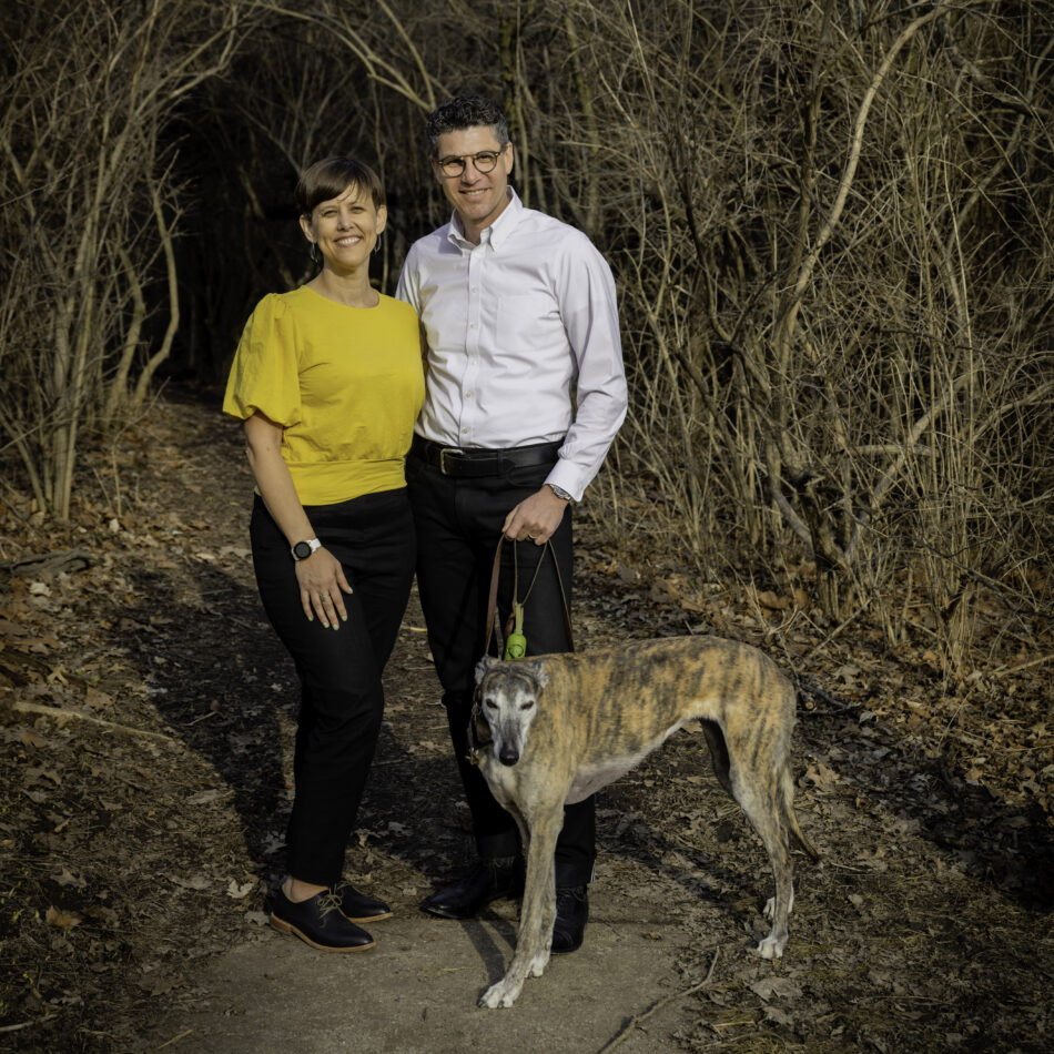 Dharma (left) and Devon (right) smiling and standing outdoors on a wooded path with their brindle greyhound, dressed casually in a yellow top and white shirt.