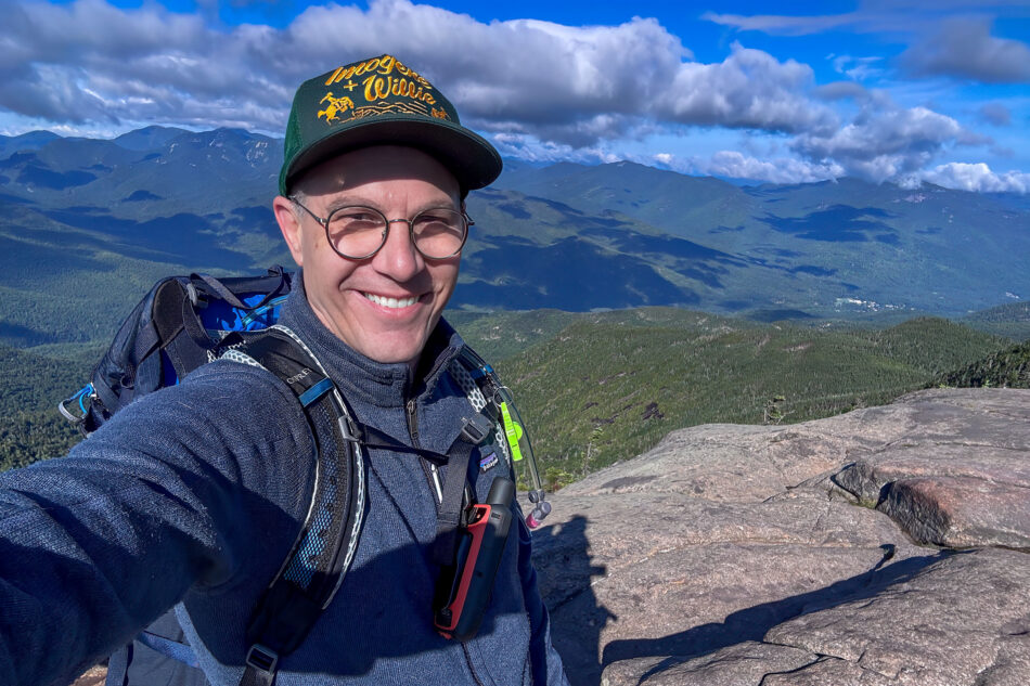 A smiling man in glasses and a trucker hat takes a selfie on a rocky summit, wearing a hiking backpack with a GPS device clipped to the chest strap, with a sweeping view of forested Adirondack peaks under a partly cloudy sky.