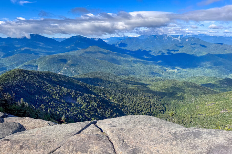 A panoramic view from a rocky summit overlooking layered green mountain ridges and a distant valley under a partly cloudy sky.