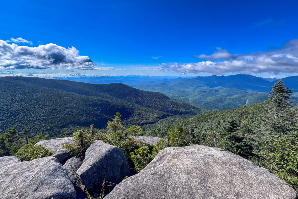 A wide view from a boulder-strewn summit looking out over forested Adirondack ridgelines toward a hazy mountain horizon.