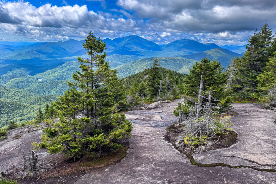 A rocky, open summit path lined with evergreen trees, overlooking layered mountain ridges under a partly cloudy sky.