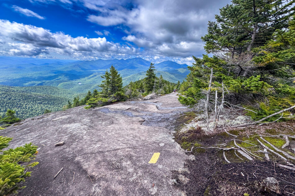 A wide slab of open rock on a mountain summit, marked with a yellow trail blaze, framed by evergreen trees and distant peaks.