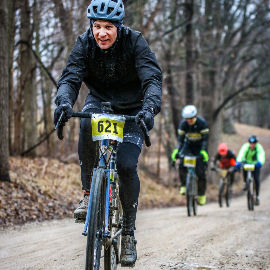 Devon Akmon in black gear and helmet, riding a gravel bike with race number 621, on a muddy dirt road during a race, with several other cyclists following in the background.