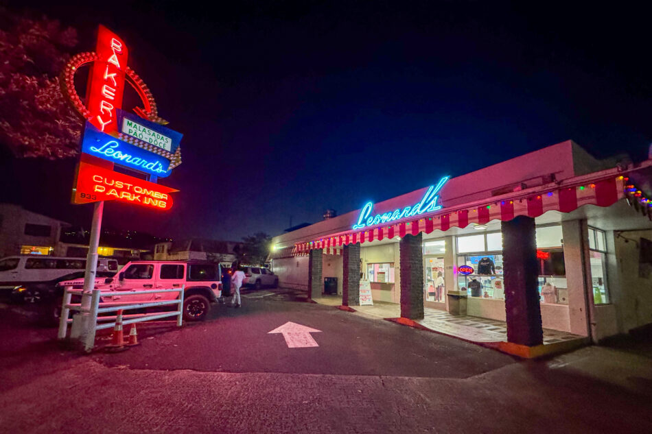 A view of Leonard's Bakery in Honolulu, Hawaii, at dawn featuring its iconic neon signs and retro-style building, with parked vehicles and a few people near the entrance.