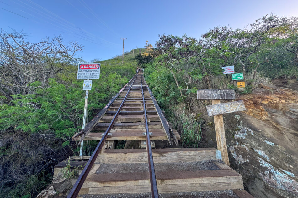 View of a warning sign on the Koko Crater Trail cautioning hikers about the unsafe railway bridge, with lush vegetation and the steep path visible ahead.