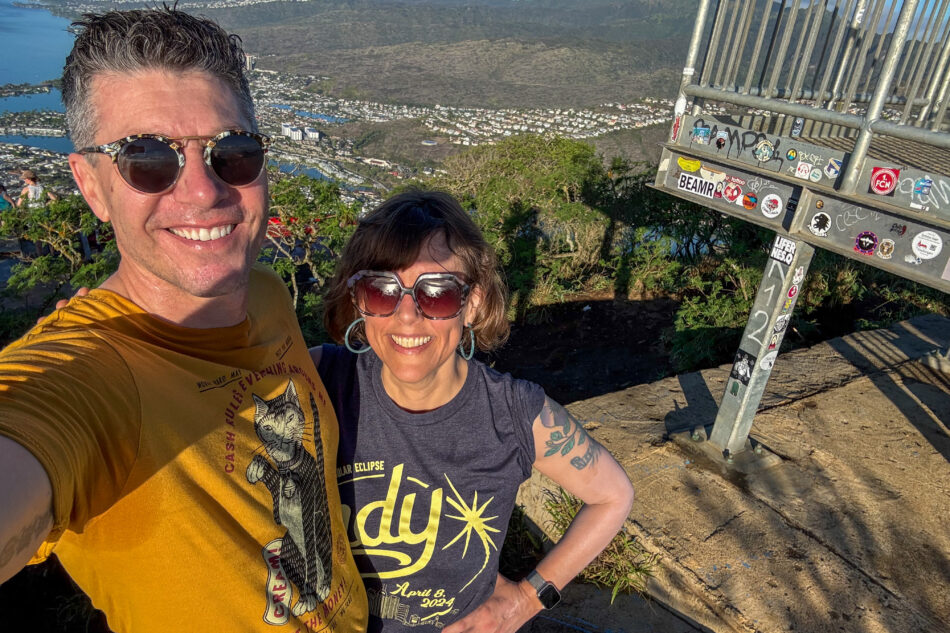 Dharma and I smiling at the summit of Koko Crater Trail, with a panoramic view of Oahu's landscape, including residential areas and water bodies, in the background.