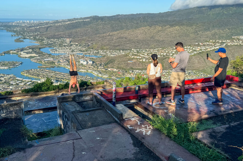 Visitors at the summit of Koko Crater Trail enjoying the stunning view of Oahu, with one person performing a handstand near the edge of the platform.