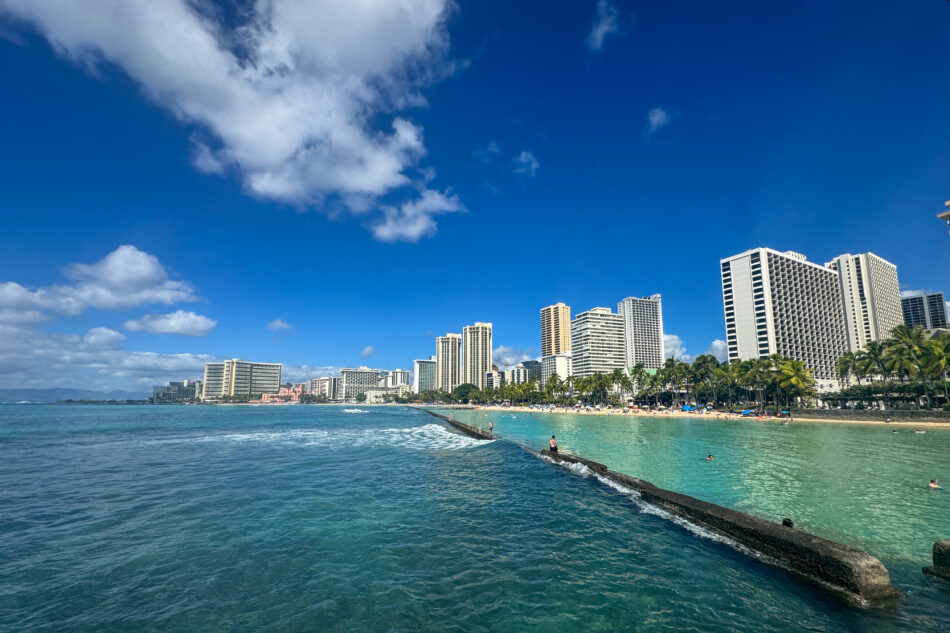 A panoramic view of Waikiki Beach, showcasing its turquoise waters, sandy shores, and a skyline of high-rise hotels and resorts under a clear, sunny sky.