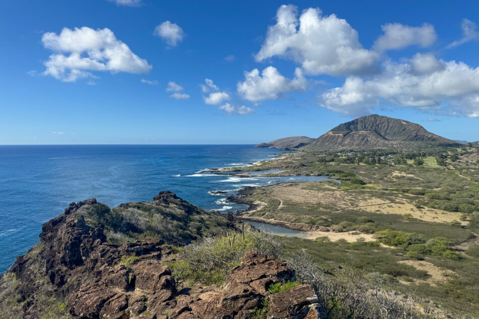 A stunning coastal view from the Makapuʻu Point Lighthouse Trail, featuring rocky cliffs, lush greenery, and the vast blue ocean under a sunny sky with scattered clouds.