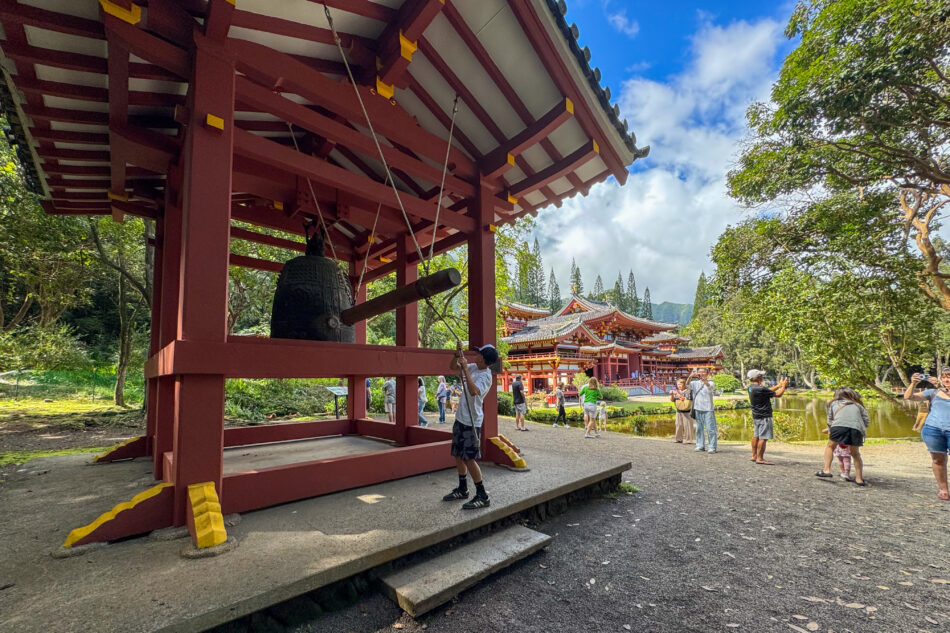 My nephew striking a large ceremonial bell at the Byodo-In Temple, with the striking red temple structure and tranquil pond surrounded by trees in the background.