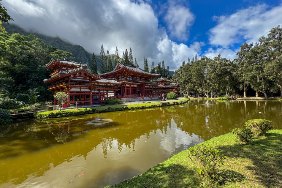 The serene Byodo-In Temple in Hawaii, reflected in a peaceful pond, with lush green landscaping and mountains shrouded in mist in the distance.