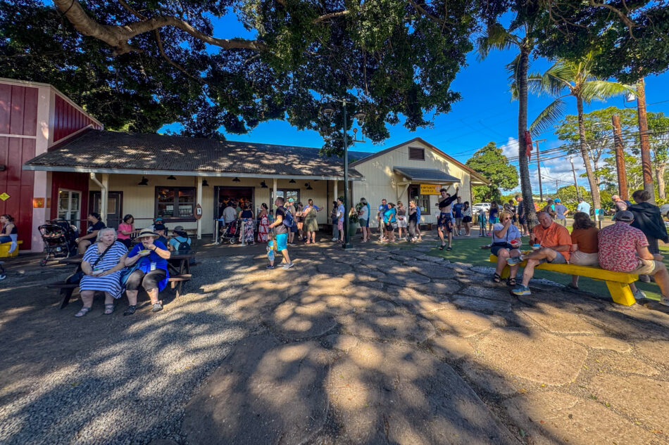 Daytime view of Matsumoto Shave Ice in Haleʻiwa, Hawaii, with a bustling crowd of visitors enjoying shave ice and relaxing under the shade of large trees, creating a lively and inviting atmosphere.