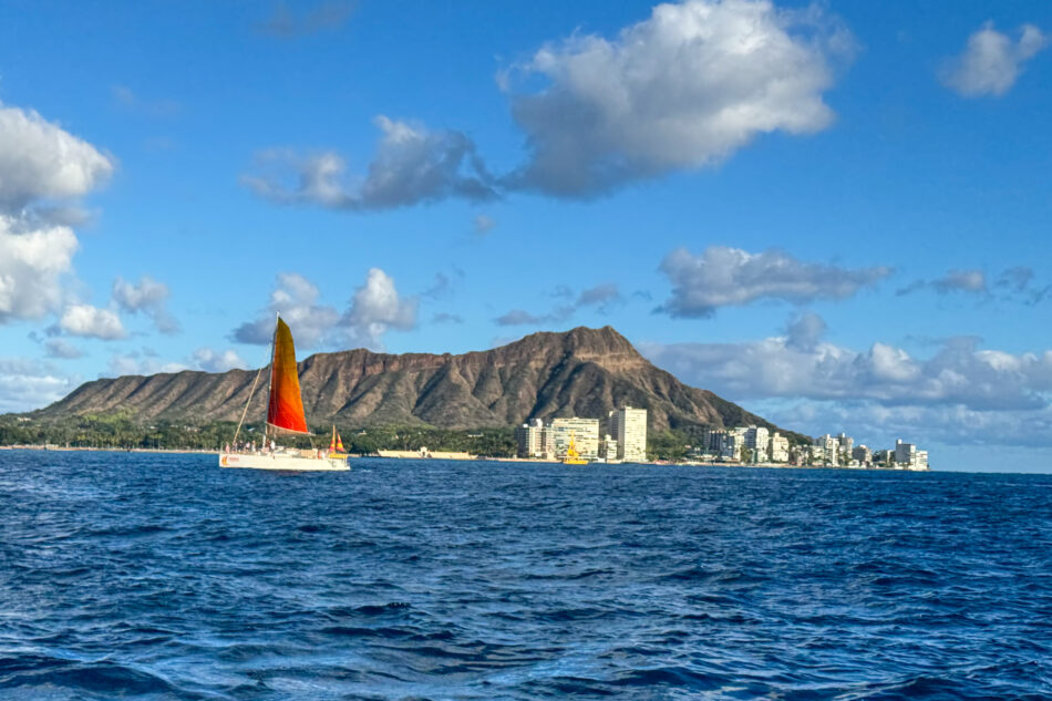 A vibrant catamaran with colorful sails gliding on the blue waters near Waikiki, with the iconic Diamond Head crater and city skyline in the background.