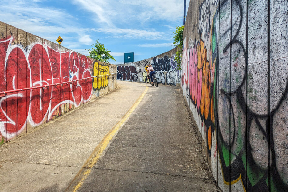 Dharma riding along the Lachine Canal bike path bordered by high walls covered in vibrant graffiti under a clear blue sky.