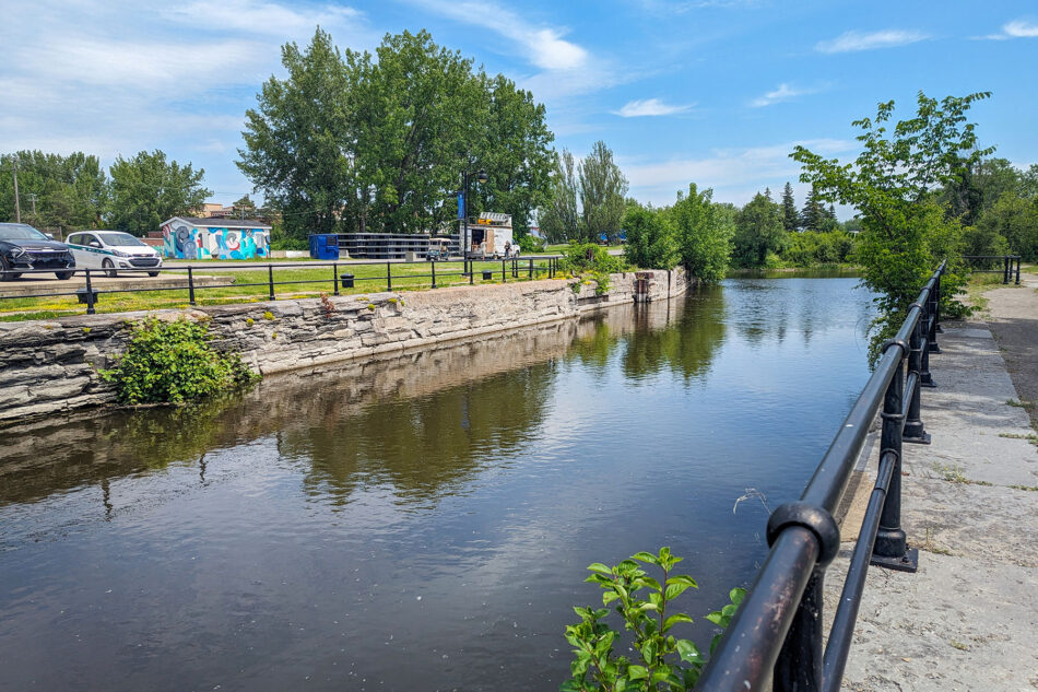 Scenic view of a Lachine Canal with a mural-painted building on one side and a lush green pathway on the other, under a clear blue sky.