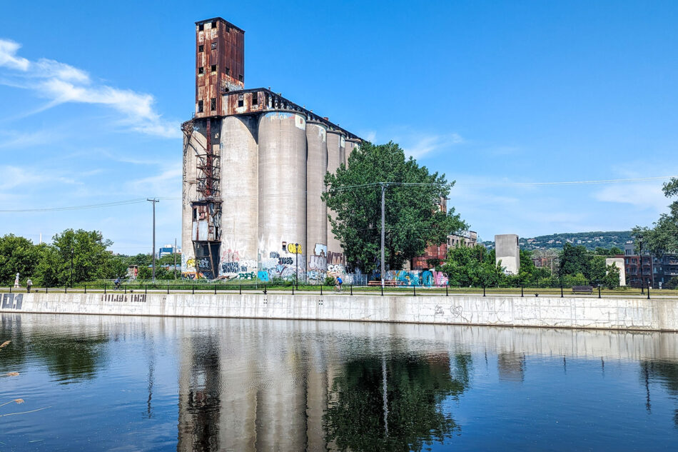 Large, rusted grain silos covered in graffiti reflecting on the calm water of the Lachine Canal, under a sunny sky.