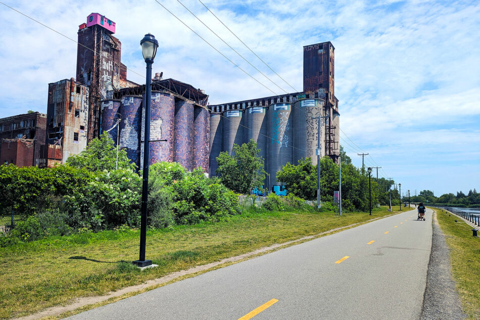 Old industrial silos covered in colorful graffiti and a vibrant pink top, located beside a green park and bike path under a sunny sky.