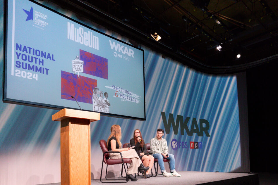 A panel discussion at the 2024 National Youth Summit on Elections and Politics. Three panelists, including two young people, sit on stage in front of a large screen showing the summit's logo and graphics. The stage is branded with the logos of the National Museum of American History, WKAR, PBS, and NPR. One panelist is holding a microphone, engaged in conversation.