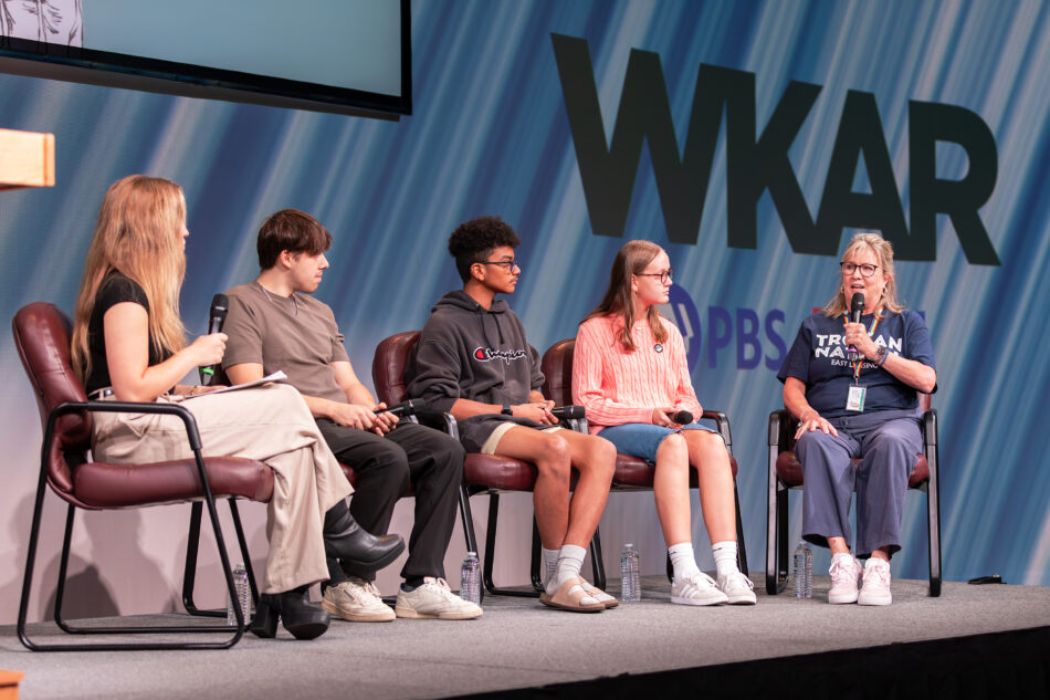 A diverse panel of four people, including three young individuals and one older adult, sits on stage during a discussion at the 2024 National Youth Summit. The participants hold microphones, and the backdrop features the logos of WKAR, PBS, and NPR. The adult panelist is speaking while the younger participants listen attentively.