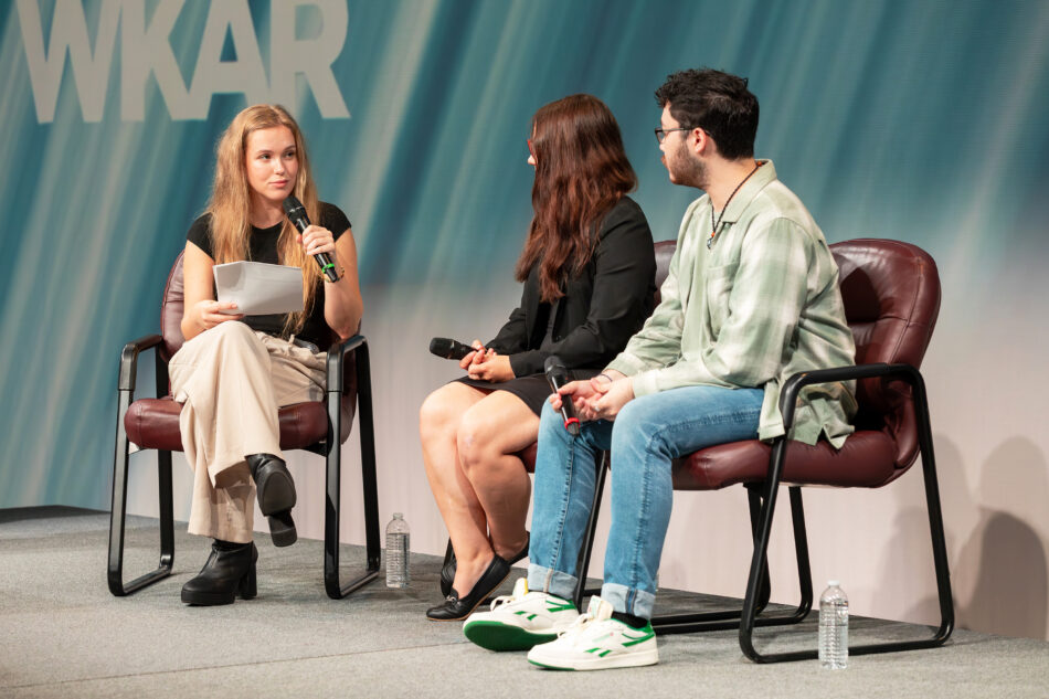 A panel featuring three individuals is seated on stage during the 2024 National Youth Summit. The moderator holds a notepad and microphone while leading the discussion. The two other panelists, a young woman and a young man, listen and prepare to speak. WKAR and PBS logos are displayed prominently on the backdrop.