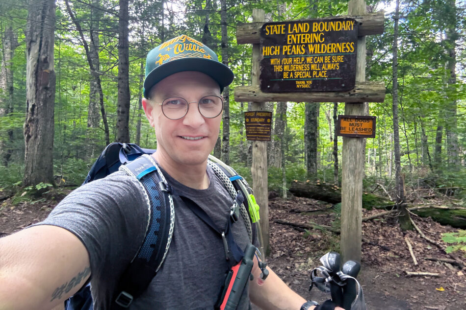 A smiling man in glasses and a cap poses with trekking poles beside a wooden trailhead sign reading 'State Land Boundary, Entering High Peaks Wilderness,' in a green forest.