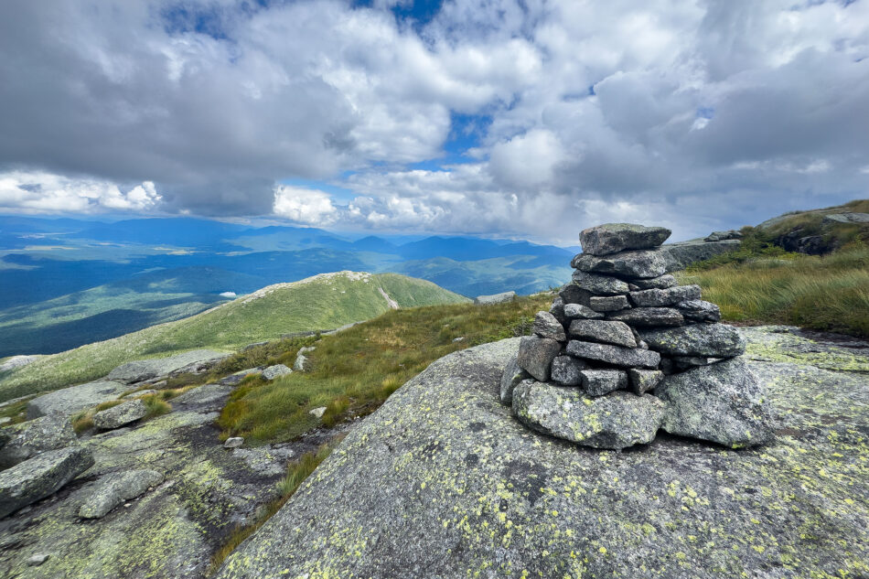 A stone cairn marks an open, grassy alpine summit with a sweeping view of layered green mountains and a partly cloudy sky.
