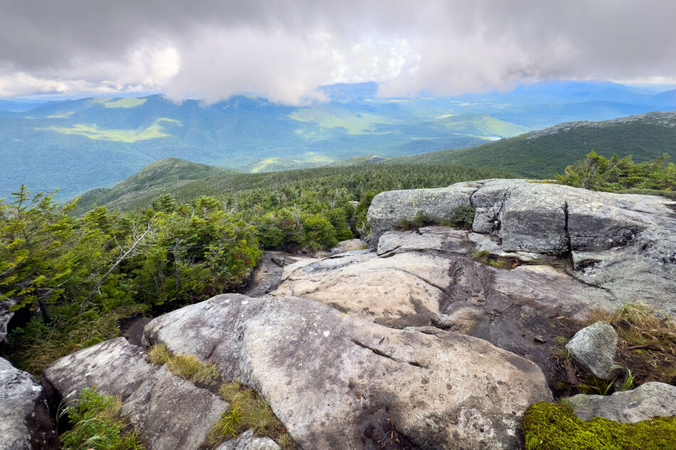 wide rock ledge on an alpine summit overlooks forested mountains fading into a hazy, cloud-filled horizon.