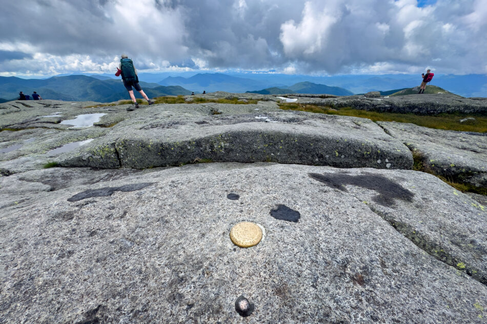 Hikers with backpacks walk across a rocky, grass-tufted alpine summit dotted with small pools, with a brass survey marker embedded in the rock in the foreground.