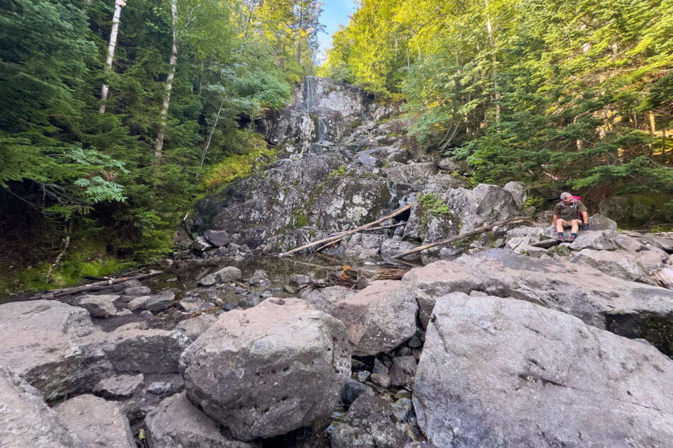 A hiker with a backpack rests on a boulder beside a small pool at the base of a rocky, moss-covered streambed in the forest.