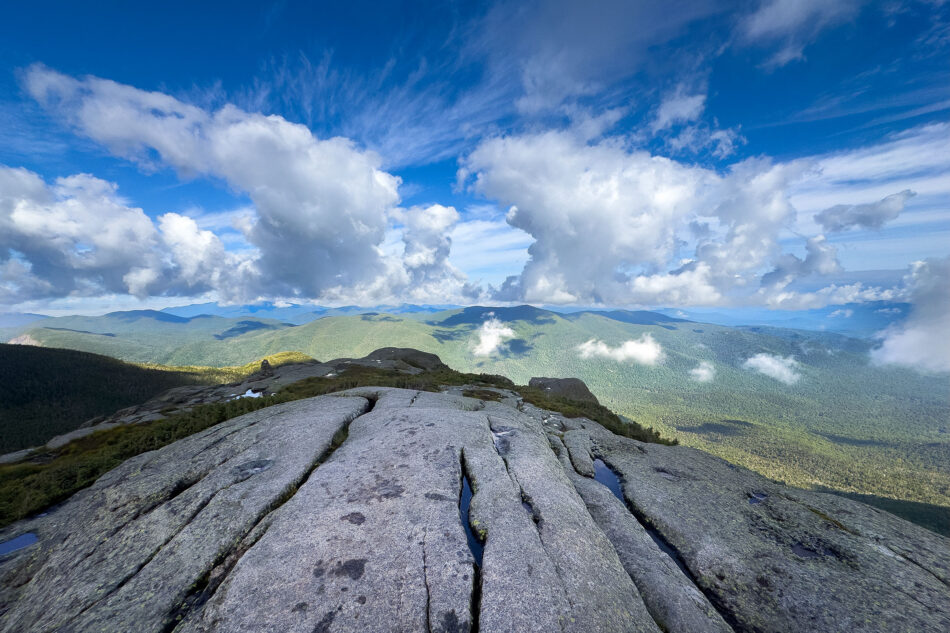 A cracked granite summit ledge overlooks forested mountains and a dramatic sky filled with tall white clouds.
