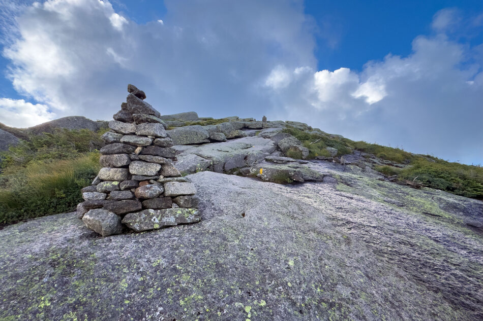 A tall stone cairn stands on a rocky alpine ridge trail, with another cairn visible in the distance under a partly cloudy sky.