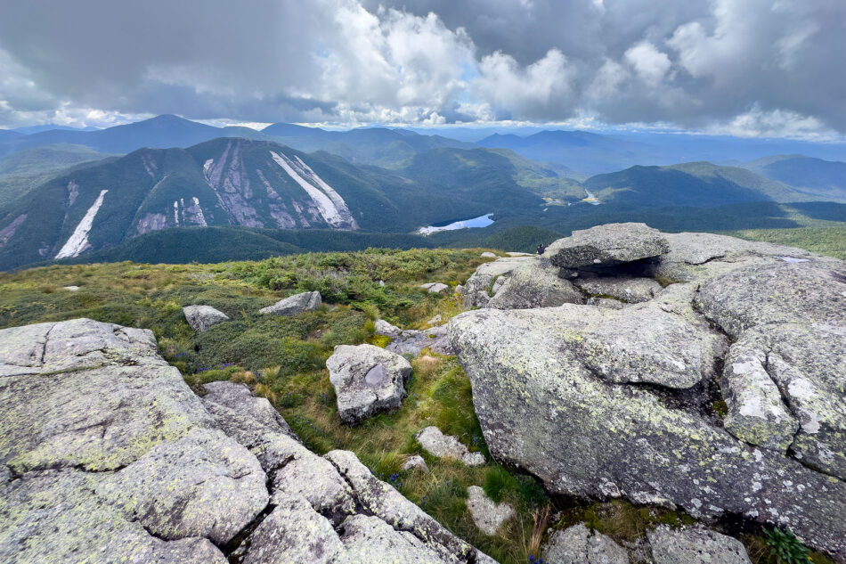 A view from a rocky summit overlooking a mountain scarred by pale slide tracks, with a small lake nestled in the forested valley below.