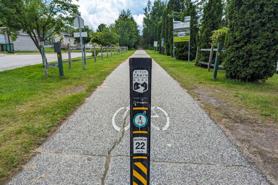 P'tit Train du Nord trail marker in a small town, with a pathway extending into the distance flanked by trees and a wooden fence.