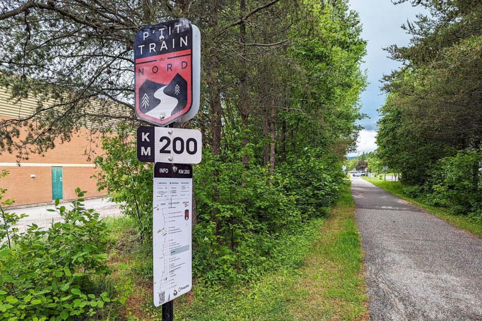 KM 200 marker on the P'tit Train du Nord trail, with a path stretching into the forested distance, flanked by greenery and a building to the side.