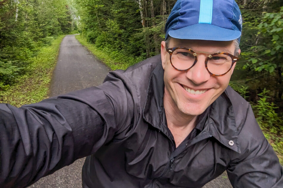 Close-up of a smiling Devon wearing glasses and a blue cap, riding on a tree-lined path.