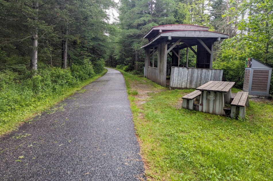 Picnic area with a wooden shelter and benches along the P'tit Train du Nord trail, surrounded by trees and greenery.