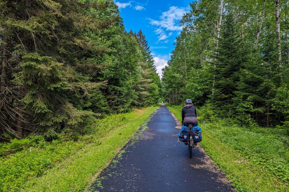 Dharma riding on a freshly paved section of the P'tit Train du Nord trail, surrounded by dense trees and greenery on a sunny day.